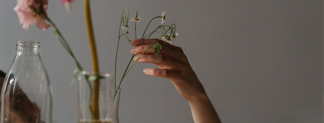 weiblich gelesene Frau schaut von links durch Glasflasche verträumt auf Blume in einer weiteren Glasflasche. Im Vordergrund steht eine weitere Glasflausche mit anderer Blume.
